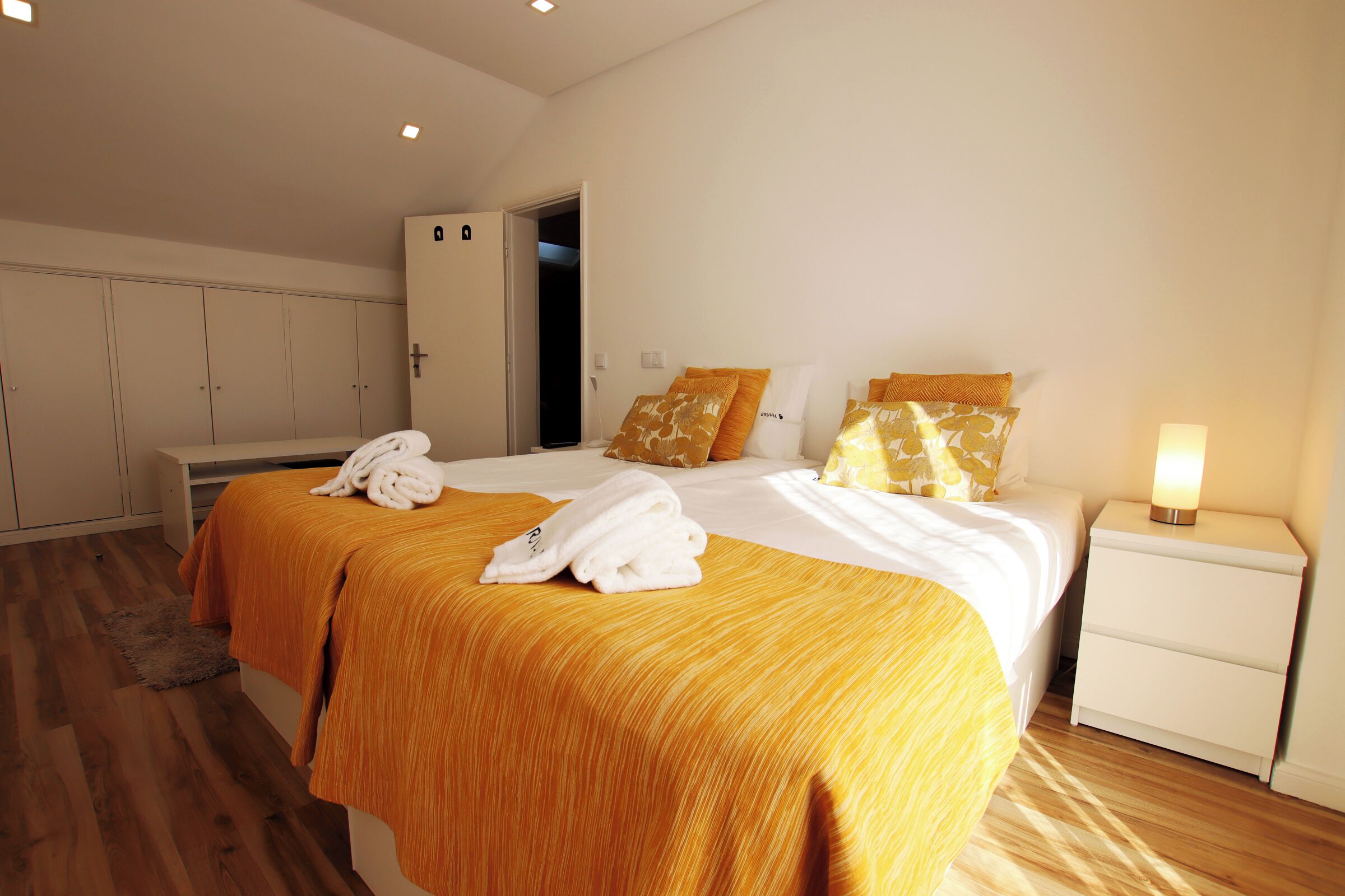 Habitación de hotel con dos camas, un armario, una mesita de noche con lámpara y una puerta al baño.