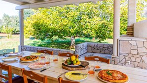 An outdoor dining area with a stone bench, a table set with plates of food, and a view of a garden.