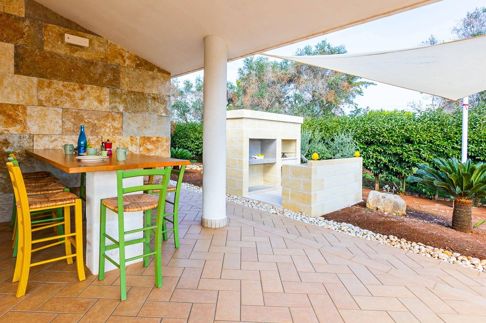 Une terrasse extérieure avec un mur en pierre, une table en bois, des chaises vertes et un espace barbecue.