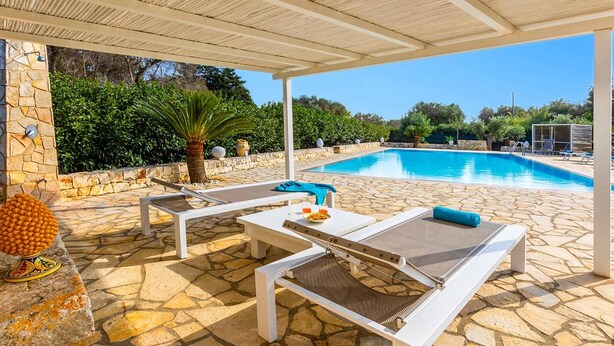 A poolside area with stone flooring, white lounge chairs, and a covered seating area.