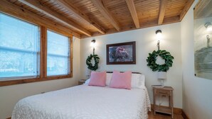 A bedroom with a wooden ceiling, a large bed with white bedding, two nightstands, a wall-mounted picture, and two wreaths.