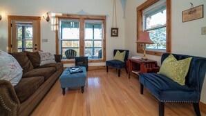 A cozy living room with a brown sofa, a blue armchair, and a small table with a lamp. There are large windows with wooden frames and a view of trees outside.