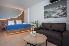 Ein modernes Hotelzimmer mit einem Bett, einer Couch, einem runden Couchtisch mit Flasche und Gläsern, einem gerahmten Bild an der Wand und einer Pflanze.