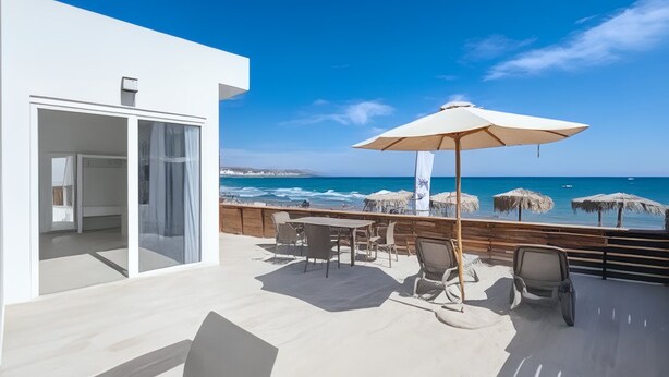 A modern outdoor terrace with a view of the ocean, featuring a table and chairs, a large umbrella, and a wooden deck. - RAFFAELLO BEACH (Rhodes)
