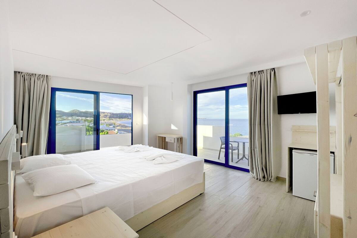 A modern hotel room with a large bed, a desk, a flat-screen TV, and a balcony with a view of the sea.