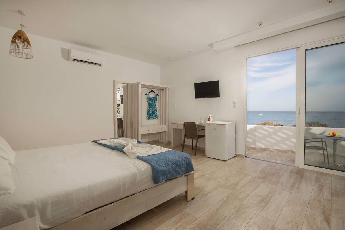 A bedroom with a bed, a desk, a chair, a TV, and a sliding glass door to the balcony.