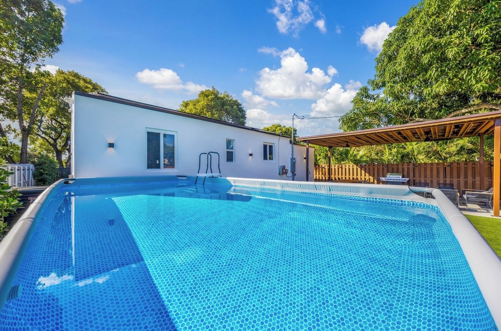 Une piscine dont le fond est recouvert de carreaux hexagonaux, un bâtiment blanc doté de fenêtres et une pergola en bois.