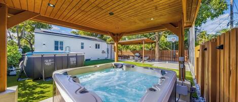 A hot tub under a wooden pergola with a backyard patio, a barbecue area, and a white house in the background.