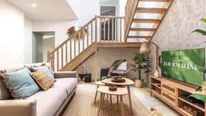 A spacious living area with a high ceiling, a wooden staircase, a sofa with cushions, a coffee table, a television, and a plant. - Cozy duplex - patio - sleeps 6 - historic village (Saint-Paul-Trois-Châteaux)
