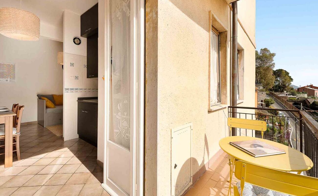 Un balcón con una mesa y una silla amarillas, una pequeña cocina con fregadero y armarios, y vistas de los edificios circundantes.