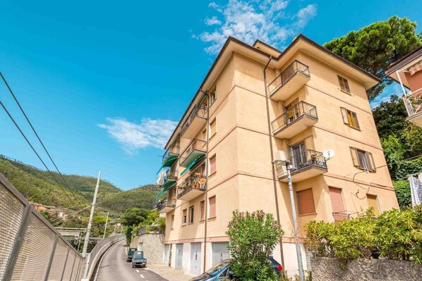 A multi-story residential building with balconies, a curved road in the background, and a hilly landscape.