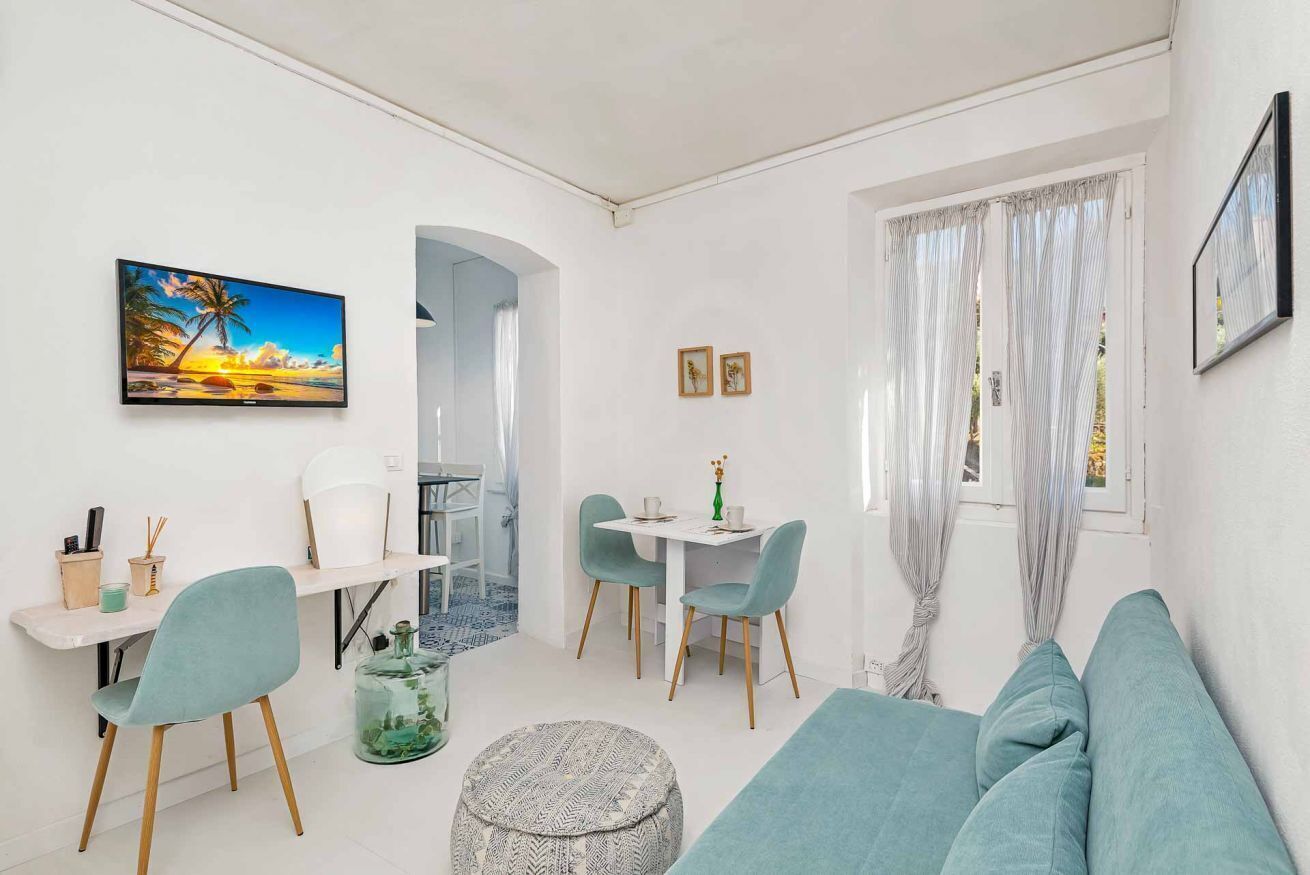 Een moderne, goed verlichte kamer met een bank, een eettafel met stoelen, een televisie aan de muur en een raam met gordijnen.