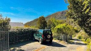 A green SUV parked on a gravel path beside a metal fence, with a mountainous backdrop and a clear blue sky. - Sea Forest - Ginko, Sea Lands (Bonassola)