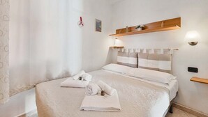 Un lit bien fait, avec du linge de lit et des serviettes blanches, une étagère en bois au-dessus et une lampe fixée au mur.