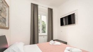 Hotelkamer met een bed, een raam met gordijnen, een aan de muur bevestigde televisie en een klein tafeltje met handdoeken.