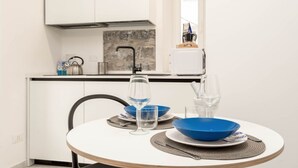 Een kleine, ronde eettafel met een witte blad en een houten onderstel, gedekt met blauwe kommen, wijnglazen en een koffiezetapparaat op het aanrecht.
