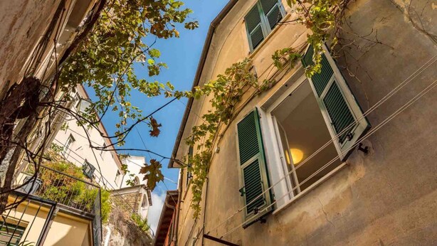 Eine schmale Straße, gesäumt von Gebäuden, darunter ein Haus mit einem Balkon und grünen Fensterläden.