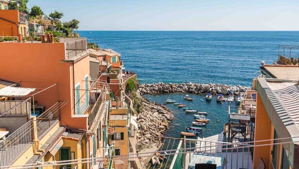 A coastal town with buildings along the sea, rocky breakwater, and boats anchored near the shore. - Enchantment, TerreMarine (Riomaggiore)