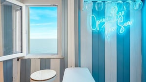 Una stanza con una grande finestra vista mare, un neon con la scritta “Open Bar The Sea” e un piccolo tavolo rotondo.