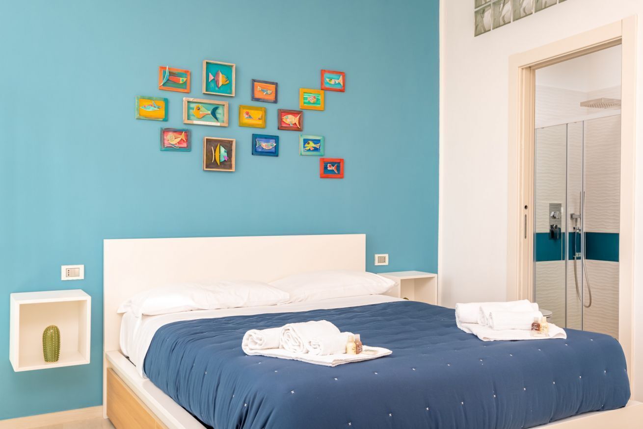 Une chambre d’hôtel avec un lit, un mur décoré de tableaux représentant des poissons aux couleurs vives, une table de chevet blanche et une salle de bain avec douche.