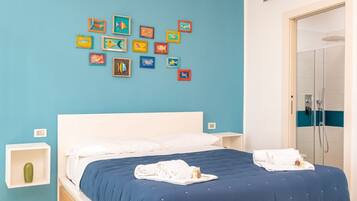 Une chambre d’hôtel avec un lit, un mur décoré de tableaux représentant des poissons aux couleurs vives, une table de chevet blanche et une salle de bain avec douche.