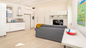 Een moderne woonkamer met een grijze bank, een klein tafeltje met een schaal, en een keuken met witte kasten en een fornuis.