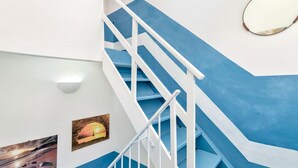 Un escalier avec des rampes blanches et des marches bleues, un luminaire fixé au mur et trois tableaux encadrés.