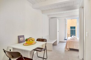 Een kleine kamer met een wit bureau, een stoel, een bed met een beige sprei en een raam met uitzicht op een gebouw.