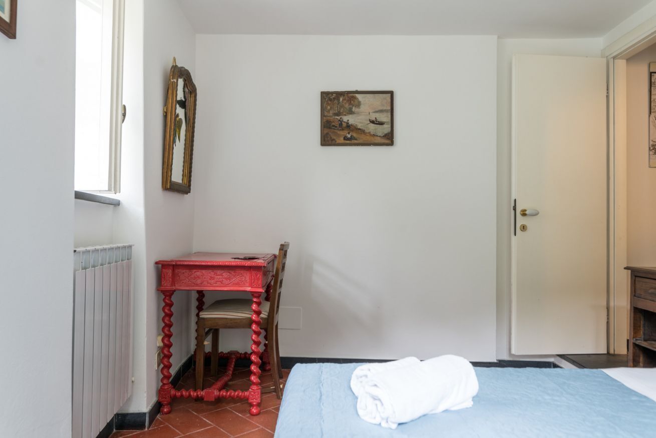 A bedroom with a bed, a red table, a chair, a painting on the wall, and a door.