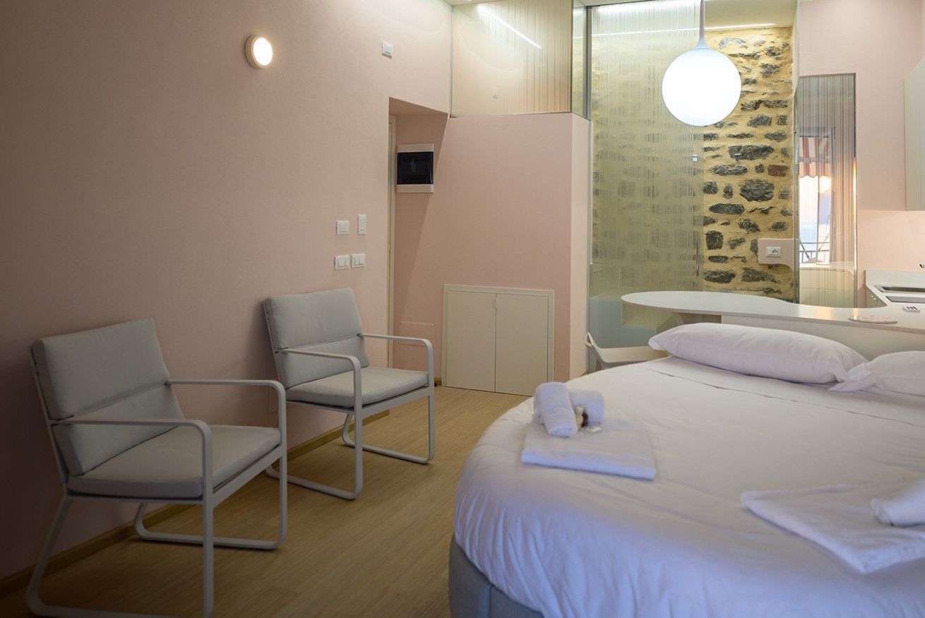 A hotel room with a bed, two chairs, a small table, a mirror, and a stone wall.