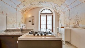 A kitchen with a central island, a sink, and a stove. There is a window with curtains and a door in the background. - The Secret Garden, TerreMarine (Lerici)