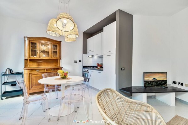 A modern kitchen with a dining area, featuring a round table, clear chairs, a wicker chair, and a TV on a black stand.