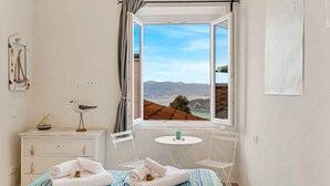 A bedroom with a bed, a bedside table, a small round table, a hanging chair, and a window with a view of a city and mountains. - La Frida, Marine Lands (Lerici)