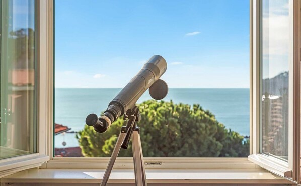 A telescope on a tripod by a window overlooking a body of water and greenery. - The Sails, Sea Lands (Lerici)