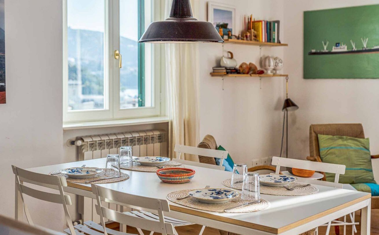A dining area with a white table set for four, a black pendant light, and a view of the outdoors through a window.