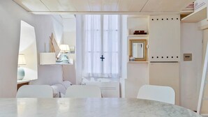 A modern, minimalist room with a marble table, white chairs, a large window with sheer curtains, and a mirror with a gold frame.