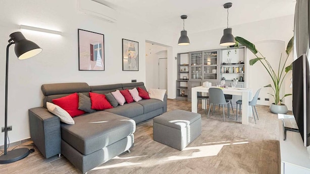 Una sala moderna con un sofá gris, un reposapiés y una zona de comedor con mesa y sillas. Hay dos lámparas colgantes, una planta grande en maceta y una estantería.