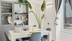 Un comedor con una mesa blanca para dos personas, una estantería con libros y adornos, y una planta grande.