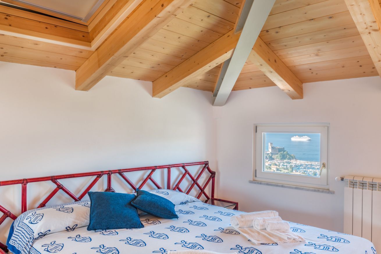 Una camera da letto con soffitto in legno, un letto con biancheria a motivi e una finestra vista su una località costiera.