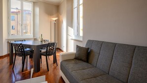 A modern living room with a grey sofa, a dining table with chairs, and large windows providing natural light.