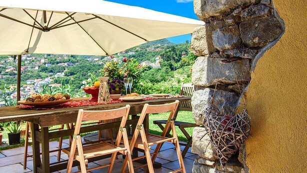 Un área de comedor al aire libre con pared de piedra, sillas de madera, una mesa dispuesta con platos y una botella, y vista a un pueblo en la ladera.