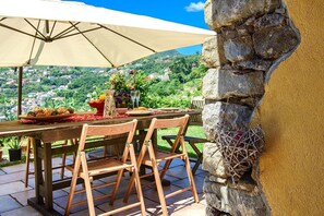 Un área de comedor al aire libre con pared de piedra, sillas de madera, una mesa dispuesta con platos y una botella, y vista a un pueblo en la ladera.