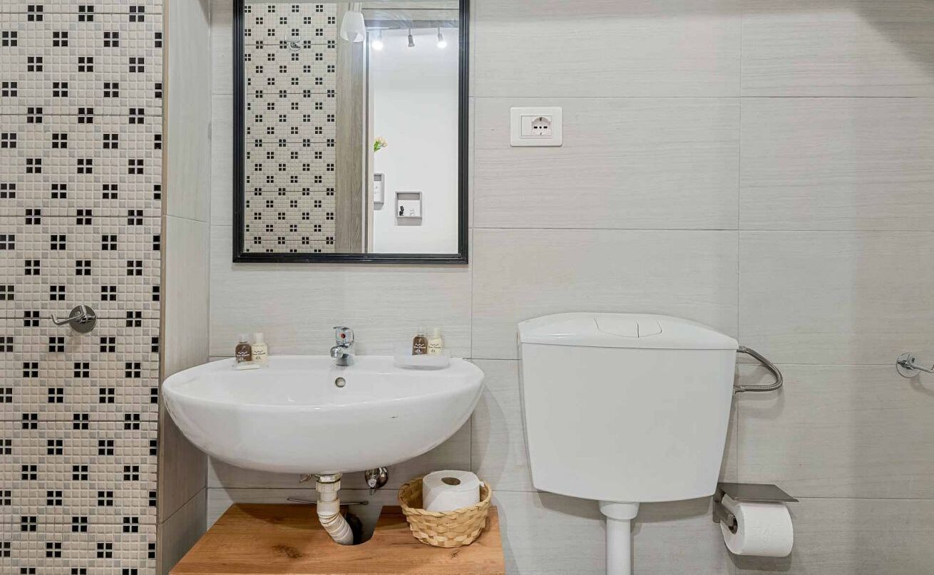A bathroom with a white sink, a toilet, and a mirror. There is a patterned tile wall on the left and a plain tiled wall on the right.