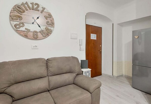 A living room with a beige sofa, a wooden clock, a door, and a refrigerator.