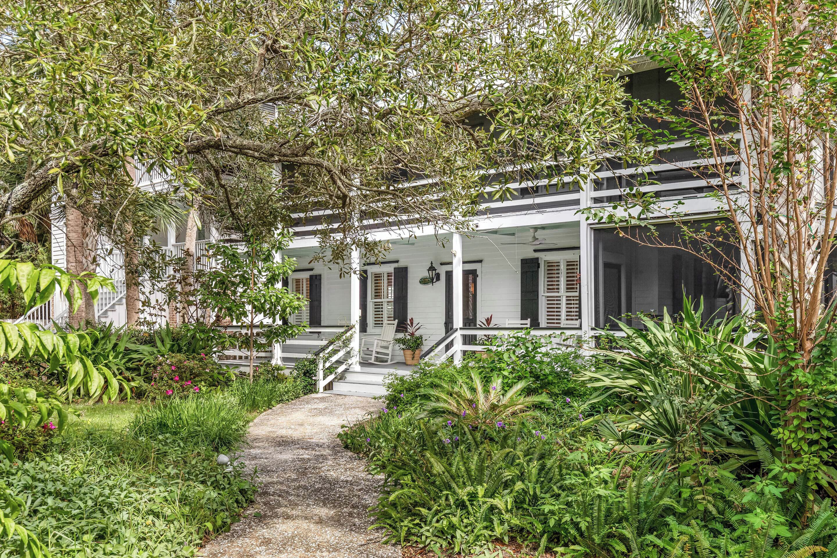 A white building with a covered porch, surrounded by lush greenery and a gravel path.