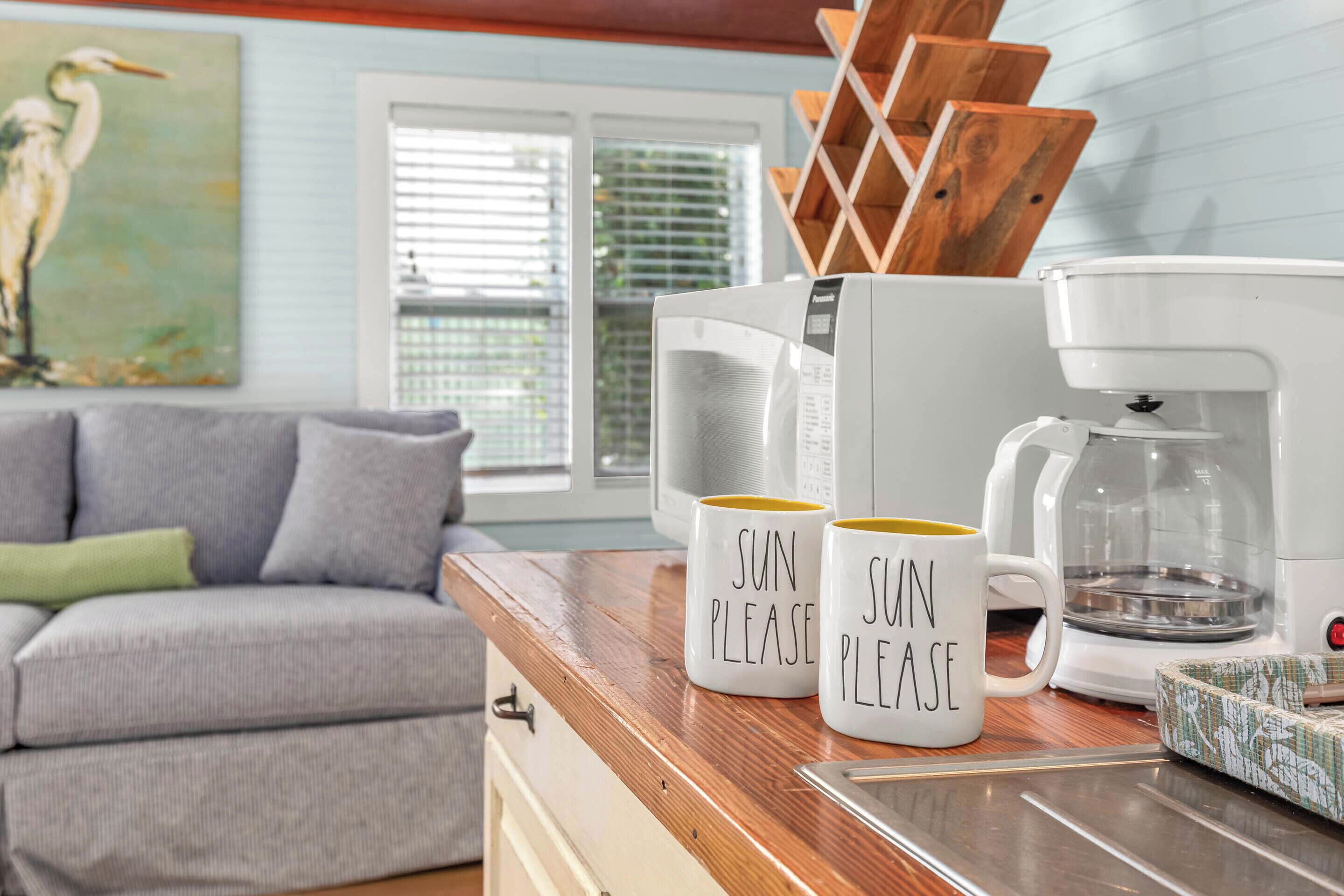 A kitchen with a coffee maker, two mugs with "SUN PLEASE" text, a wooden cutting board, and a gray couch in the background.