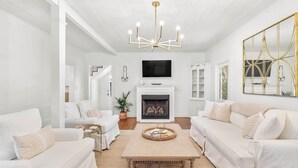 Un salon spacieux, meublé de blanc, avec une cheminée et un lustre moderne.