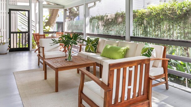 Une terrasse couverte avec des meubles en bois, un canapé blanc avec des coussins verts, et une vue sur un jardin et une maison en arrière-plan.