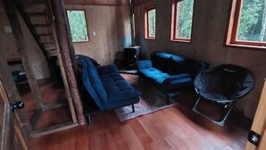Ein Blockhaus-Interieur mit einer Couch, einem Sessel, einem kleinen Tisch und einem Fenster mit Blick auf Bäume.