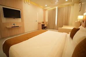 A hotel room with a large bed, a desk, a chair, a TV mounted on the wall, and a window with curtains.
